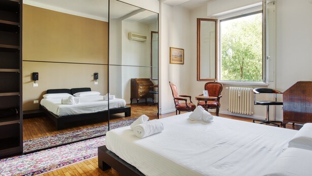A hotel room with a large bed, a desk, a chair, a mirror, and a window with a view of trees. - JOIVY Stylish Flat in Porta Genova (Milan)