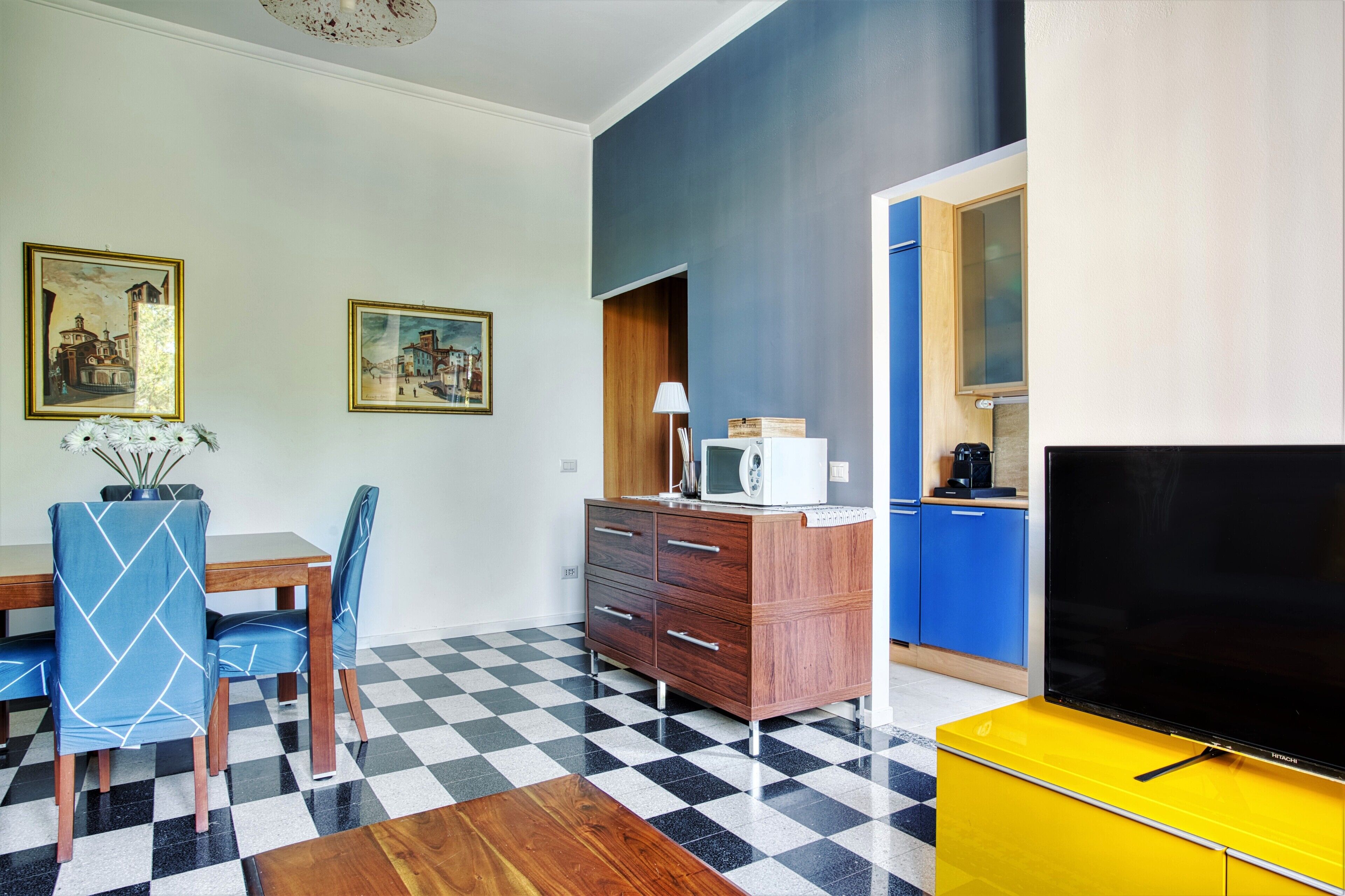 Una sala de estar moderna con piso a cuadros, un mueble de madera, un televisor sobre un soporte amarillo y dos cuadros enmarcados en la pared.