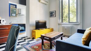 A modern living room with a checkered floor, a blue sofa, a wooden coffee table, a yellow cabinet, a television, and a window with a view of trees. - JOIVY Stylish Flat in Porta Genova (Milan)