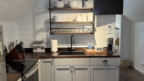Een moderne keuken met witte kastjes, een houten aanrechtblad en een zwarte kraan. Boven de gootsteen staan planken met servies en glazen.