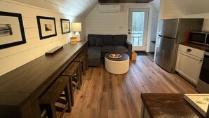 Een ruimtelijk ingerichte leefruimte met een houten vloer, een bank, een klein rond tafeltje, een kitchenette met een koelkast en magnetron, en een deur naar buiten.