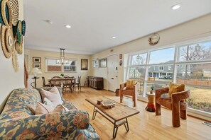 A living room with a sofa, coffee table, and two chairs, a dining area with a table and chairs, and large windows with a view of the outside.