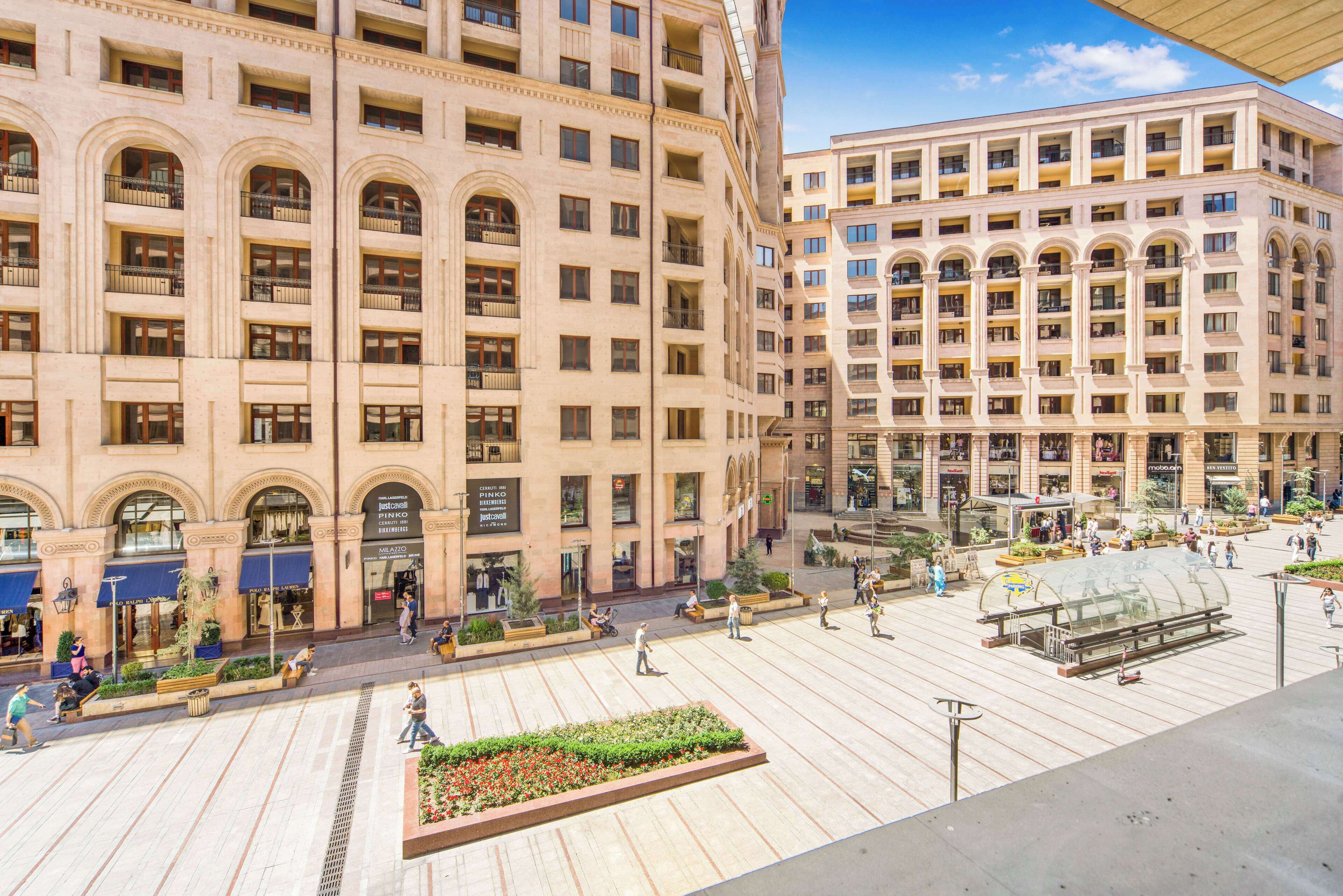 A large, multi-story hotel with a classic architectural style, featuring numerous balconies and a spacious public plaza with seating areas and greenery.