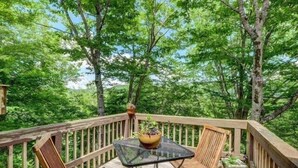 A wooden deck with a glass table and two chairs, surrounded by trees.