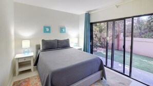 A bedroom with a bed, a nightstand, a lamp, and a view of a garden through a sliding glass door. - Modern condo: Steps from Atlantis & Beach (Bayview Lane)