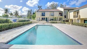 A rectangular swimming pool with a tiled edge, surrounded by a paved area with low hedges. There are two-story buildings with balconies in the background, under a clear sky. - Modern condo: Steps from Atlantis & Beach (Bayview Lane)