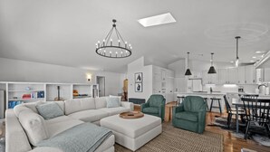 Un salon moderne avec un canapé d’angle, un coin repas avec une table et des chaises, et une cuisine dotée de meubles blancs et d’un éclairage suspendu.