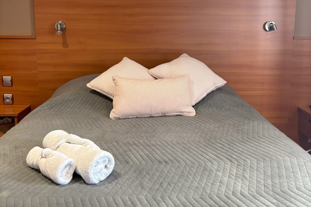 A neatly made bed with a grey quilt, two striped pillows, and rolled white towels.