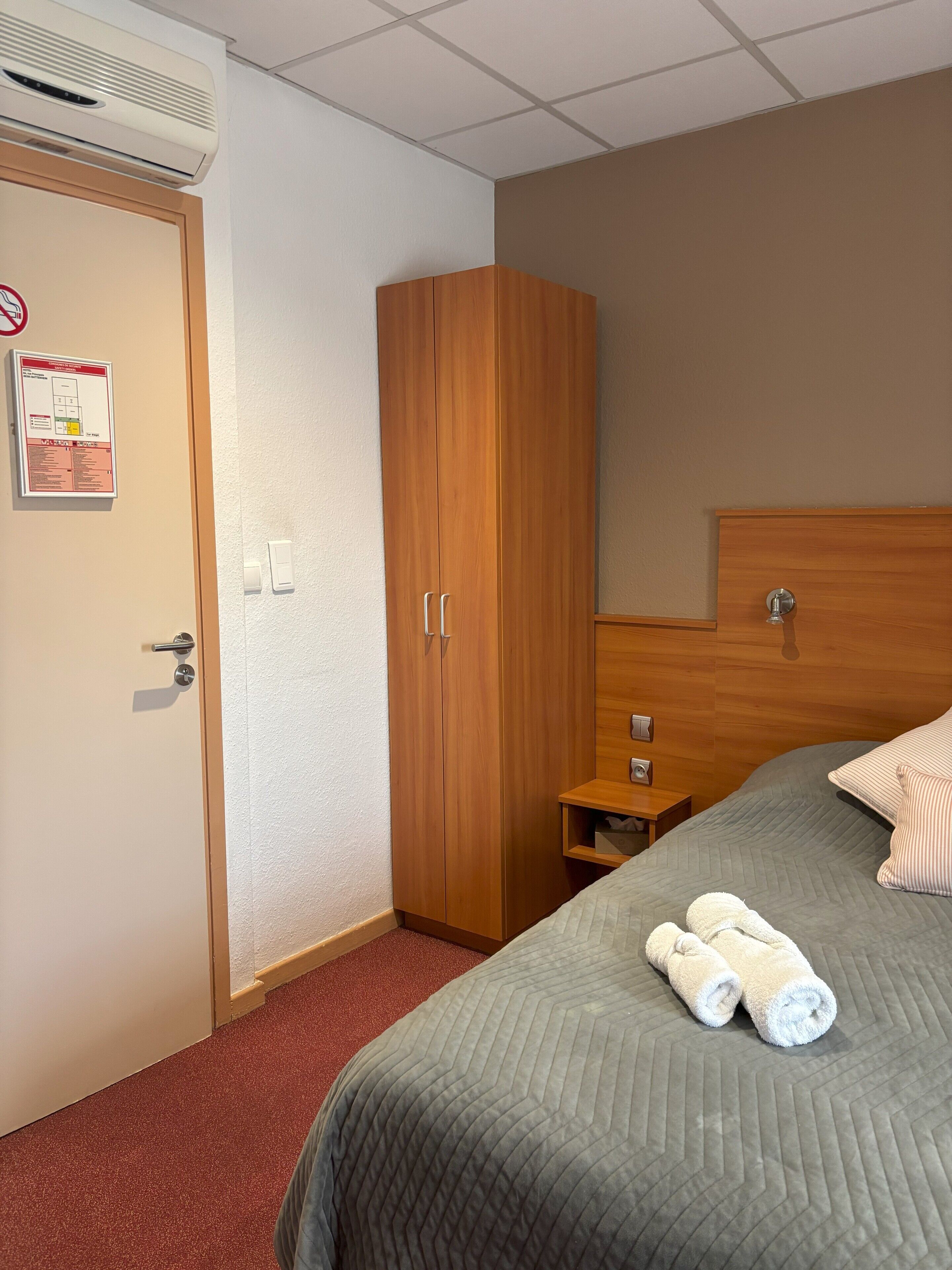 Une chambre d’hôtel équipée d’un lit, d’une armoire, d’une table de chevet et d’un climatiseur fixé au mur.