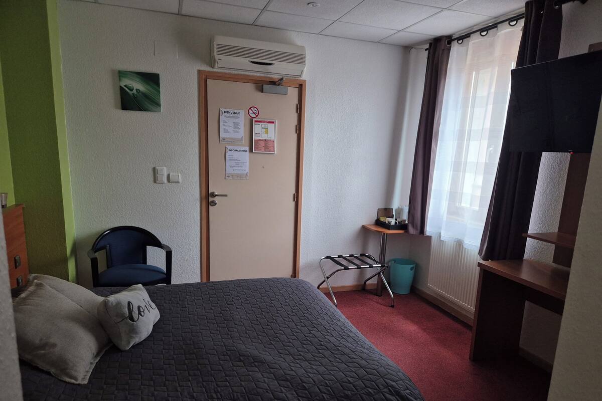 A hotel room with a bed, a chair, a desk, a waste bin, and a window with curtains.