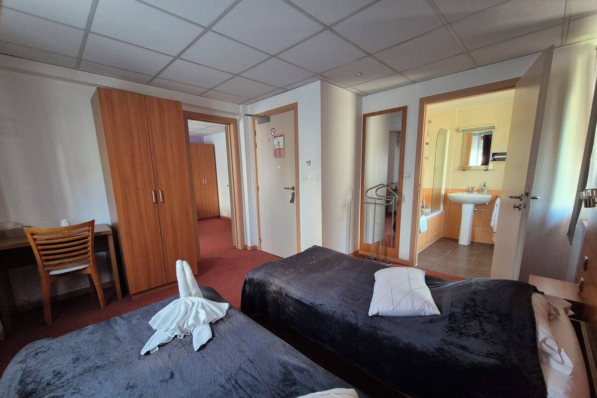 A hotel room with two beds, a desk, a chair, a wardrobe, and a bathroom visible through an open door.