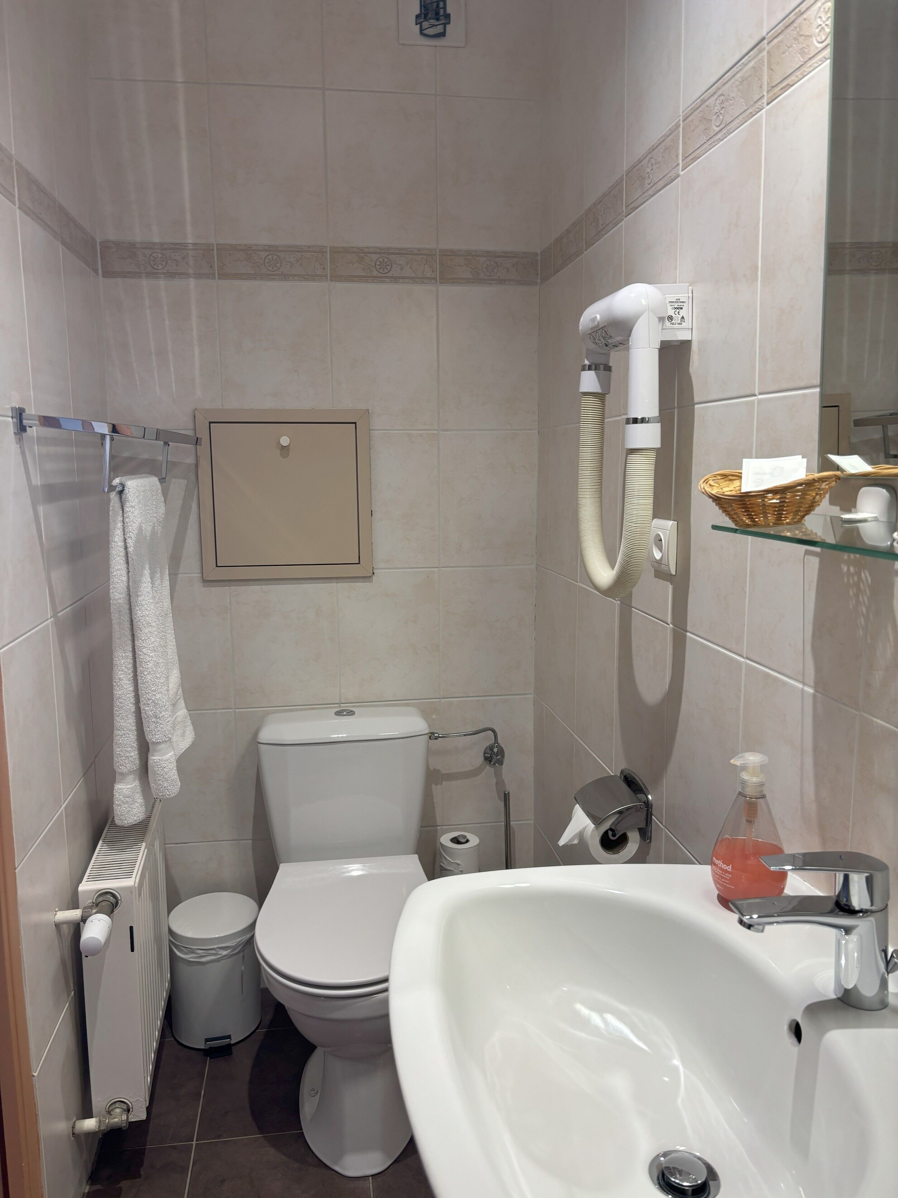 A bathroom with a toilet, bidet, and sink. There is a towel rack with towels, a waste bin, and a glass shelf with a basket and a bottle of soap.