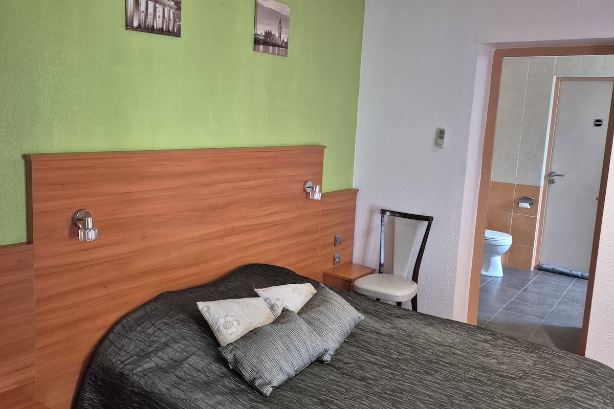 A hotel room with a wooden headboard, a large bed with a dark bedspread and pillows, a chair, and a toilet visible through an open door.