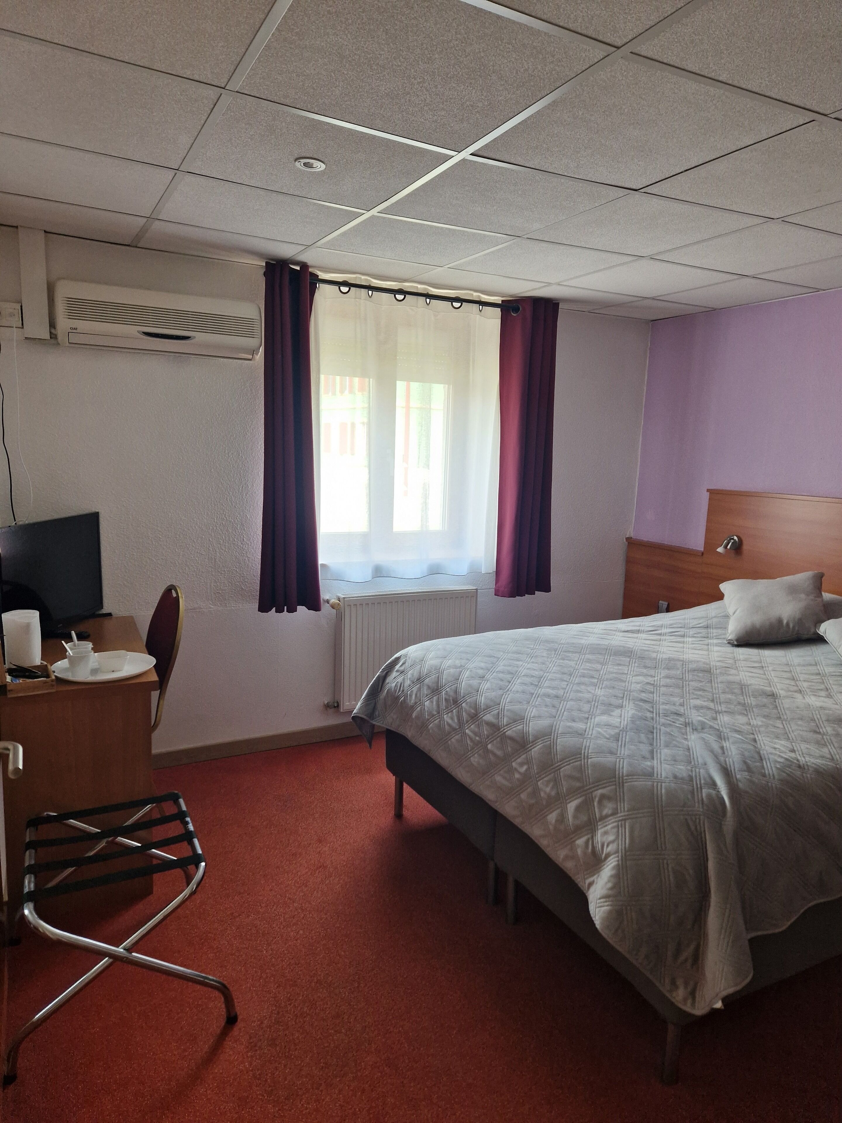 A hotel room with a bed, a desk with a chair, a radiator, and a window with curtains.