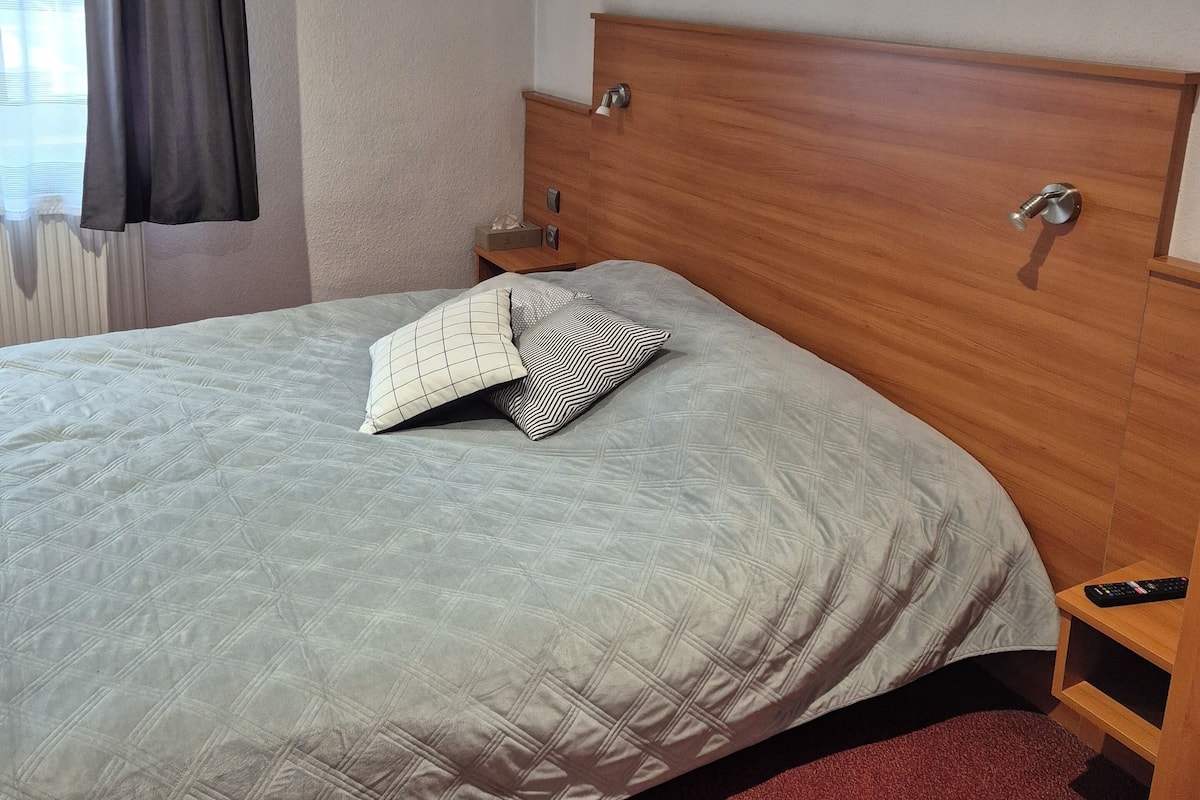 A hotel room with a bed, a nightstand, a wall-mounted picture, and a window with curtains.