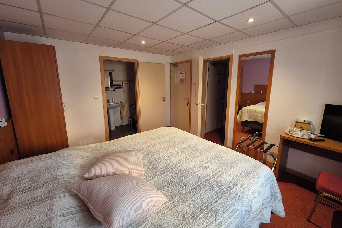 A hotel room with a bed, a desk with a chair, a TV, and a bathroom visible through an open door.