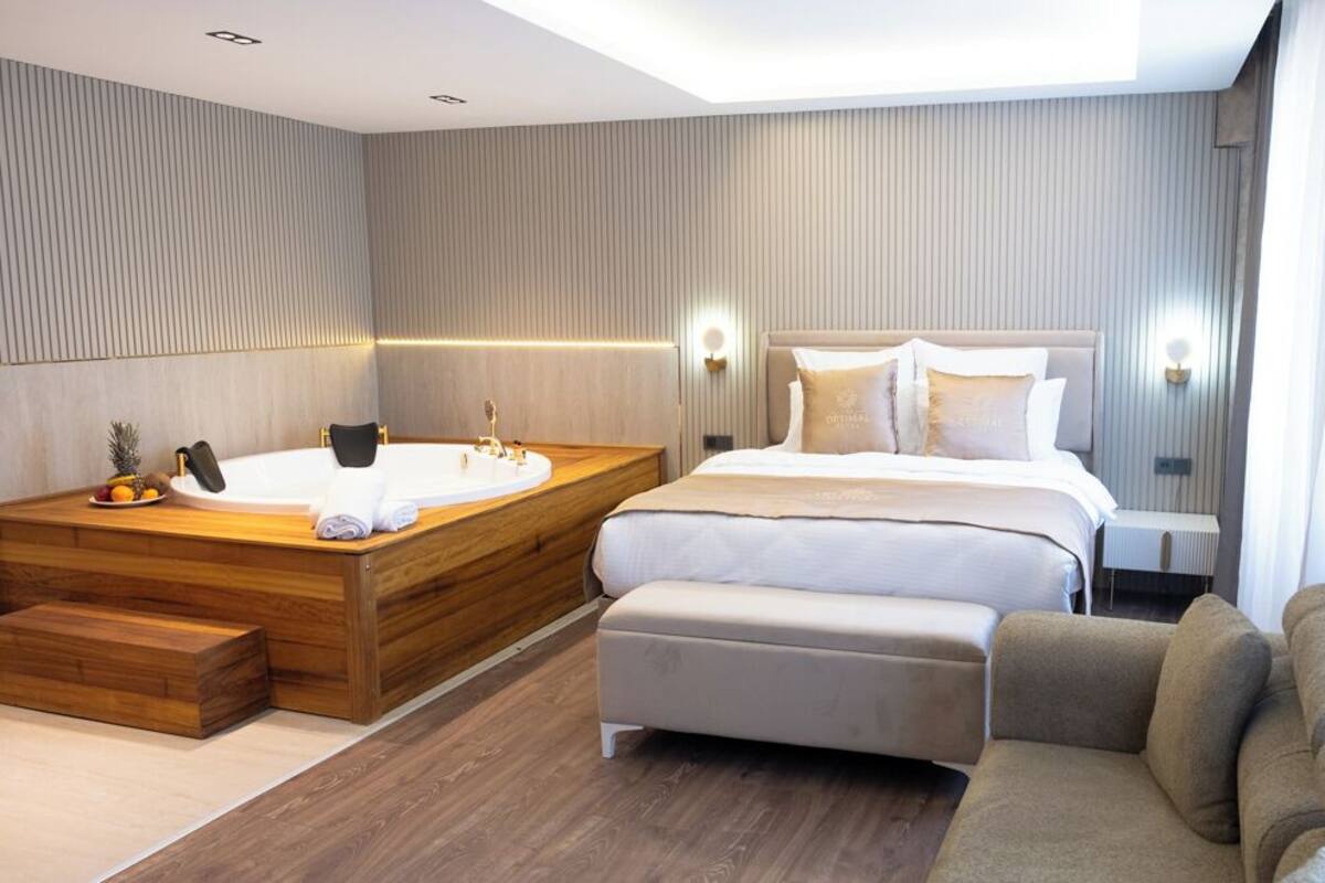 A modern hotel room with a wooden bathtub, a large bed with white linens, a sofa, and a small table.