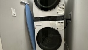 Una sala de bugaderia amb dues rentadores superposades, una taula de planxar blava i un cubell de brossa amb detergent Tide.