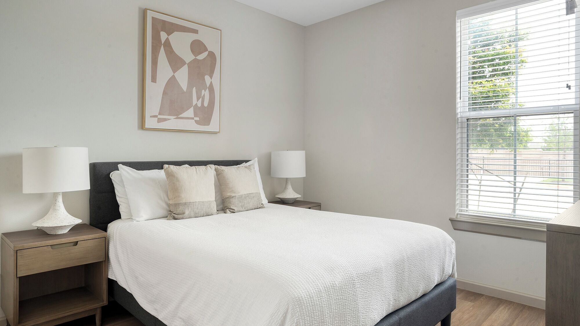 A bedroom with a bed, two bedside tables, two lamps, a window with blinds, and a wall-mounted artwork.