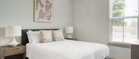A bedroom with a bed, two bedside tables, two lamps, a window with blinds, and a wall-mounted artwork.