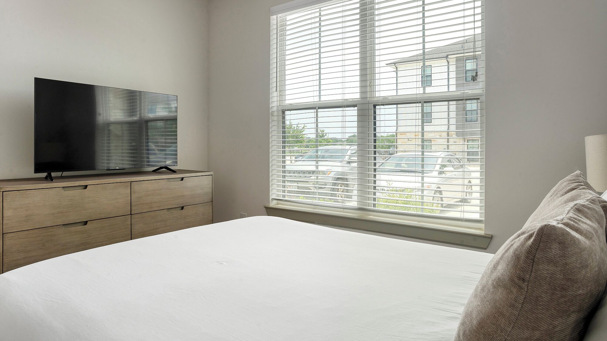 Ein Schlafzimmer mit großem Fenster, einem Fernseher auf einer Holzkommode und einem Bett mit Kissen.