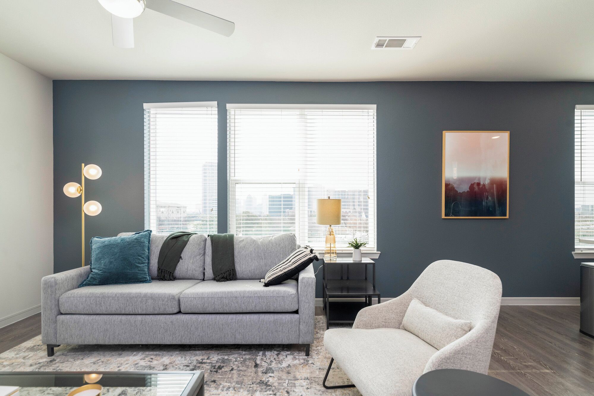 Ein Wohnzimmer mit grauem Sofa, einem weißen Sessel, einer Stehlampe und einem Bild an der Wand.