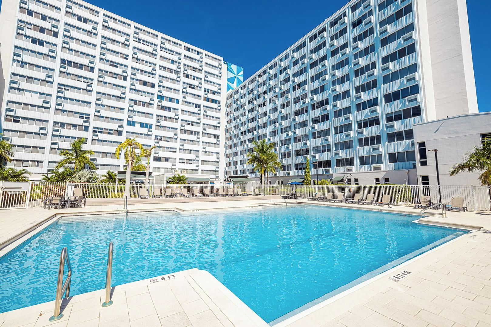 A swimming pool with a 5 ft depth marker, surrounded by lounge chairs and palm trees, with modern high-rise buildings in the background.