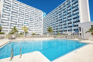 Ein Schwimmbecken mit Tiefenkennzeichnung von 1,50 Metern, umgeben von Liegestühlen und Palmen, mit modernen Hochhäusern im Hintergrund.