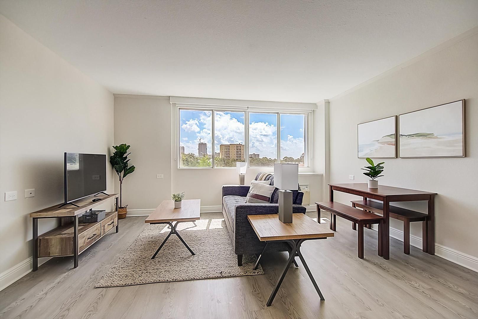 A modern living room with a flat-screen TV, a sofa, a coffee table, and a dining table. There is a large window with a view of buildings and a plant on the windowsill.