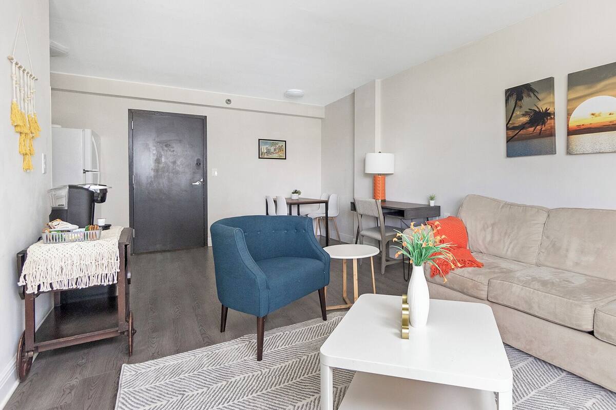Un salon avec un fauteuil bleu, un canapé beige, une table basse blanche et une petite table avec une lampe. Il y a deux tableaux au mur et une porte au fond.
