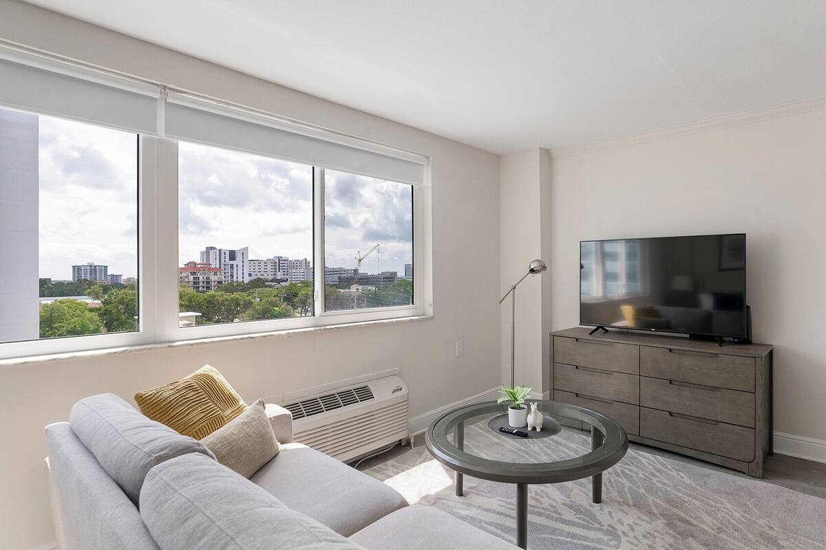 Un salon moderne avec un canapé, une table basse en verre, un téléviseur posé sur un meuble et une vue sur la ville grâce à de grandes fenêtres.