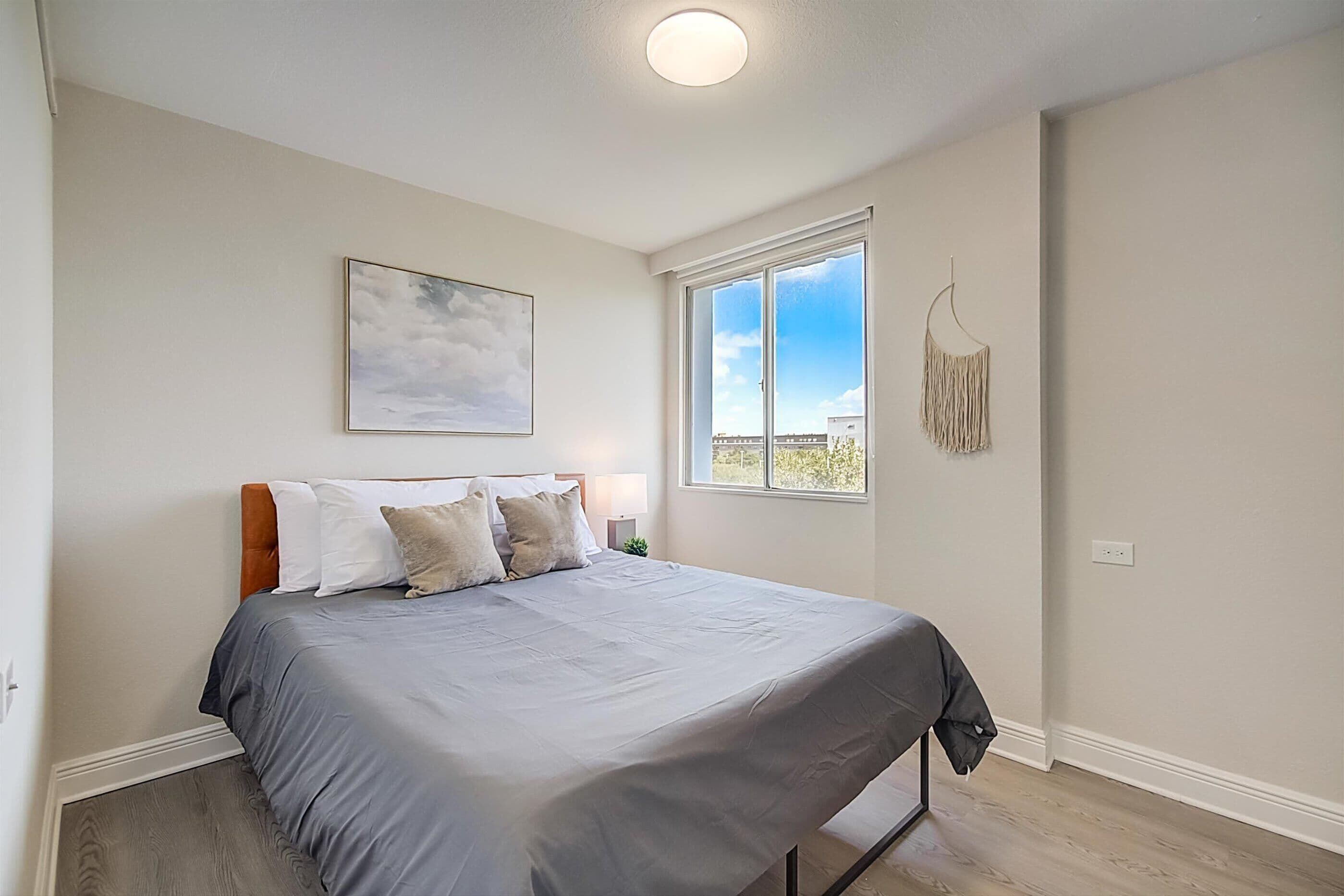 A bedroom with a bed, a window, a wall-mounted picture, and a hanging macrame piece.