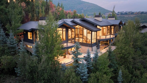 A modern multi-story house with large windows, surrounded by dense forest and hills in the background. - Luxe Estate by InvitedHome | Theater, Gym, Decks, Views, Hot Tub, Ski-In/Out (Park City)