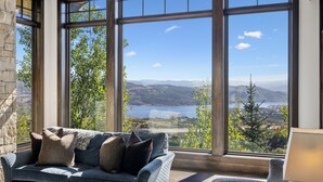 A modern living room with a large window offering a scenic view of mountains and a lake. - Luxe Estate by InvitedHome | Theater, Gym, Decks, Views, Hot Tub, Ski-In/Out (Park City)