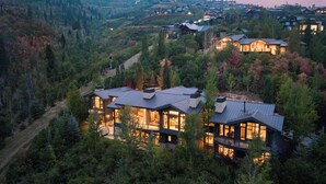 A modern, multi-story house with large windows and a metal roof, surrounded by dense forest and other residential buildings. - Luxe Estate by InvitedHome | Theater, Gym, Decks, Views, Hot Tub, Ski-In/Out (Park City)