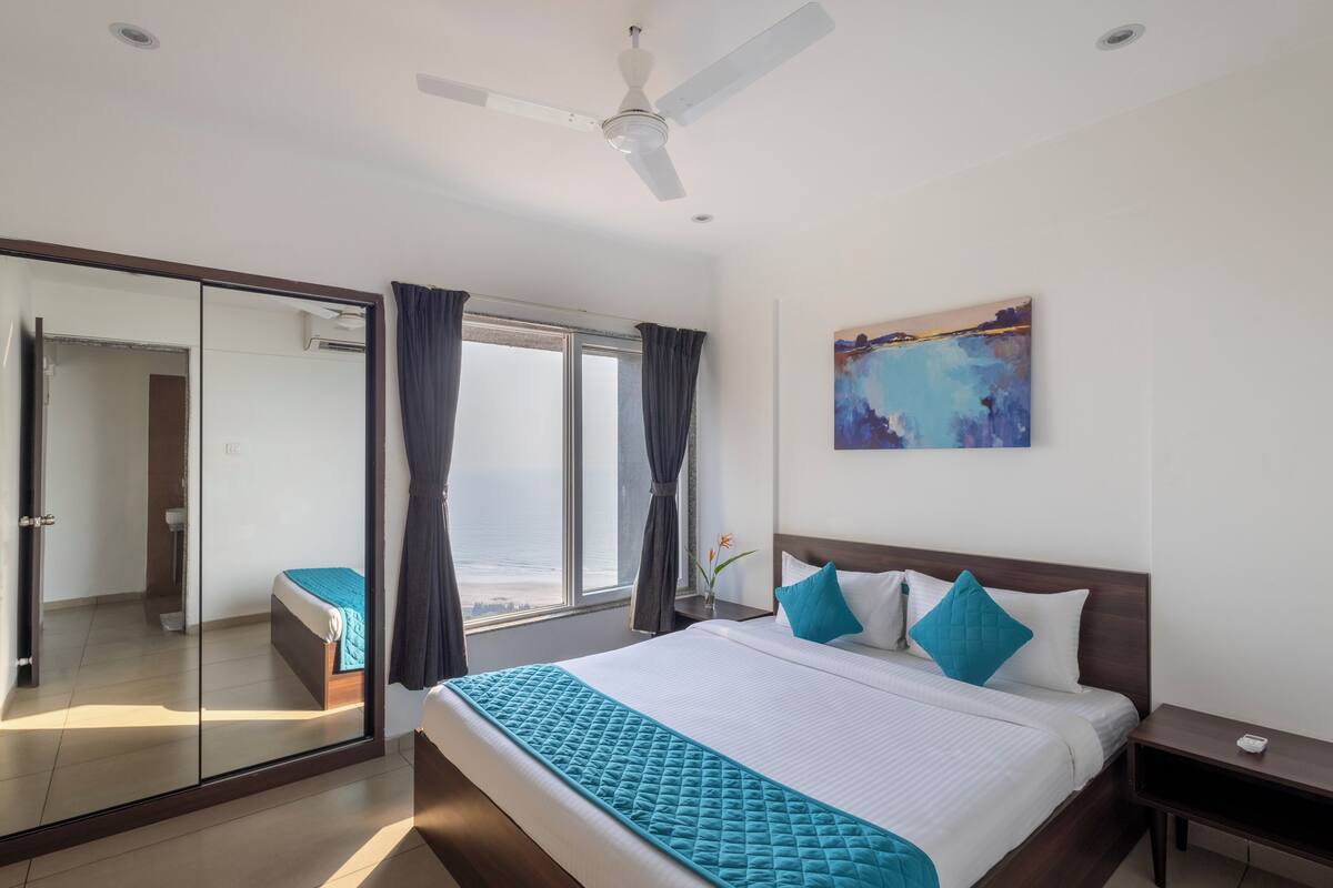 A hotel room with a bed, a ceiling fan, a window with curtains, a painting on the wall, and a nightstand.