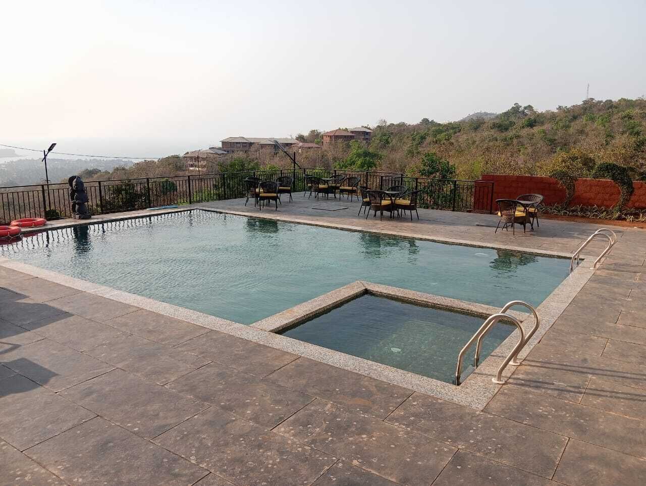Una piscina rectangular con una sección interna más pequeña, rodeada por un solárium con baldosas. Varias sillas y mesas están dispuestas alrededor de la piscina, y se observa una pared roja al fondo.