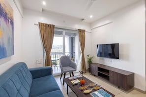 Een moderne woonkamer met een blauwe bank, een grijze fauteuil, een houten salontafel met lekkernijen, een flatscreen televisie en een balkon met gordijnen.