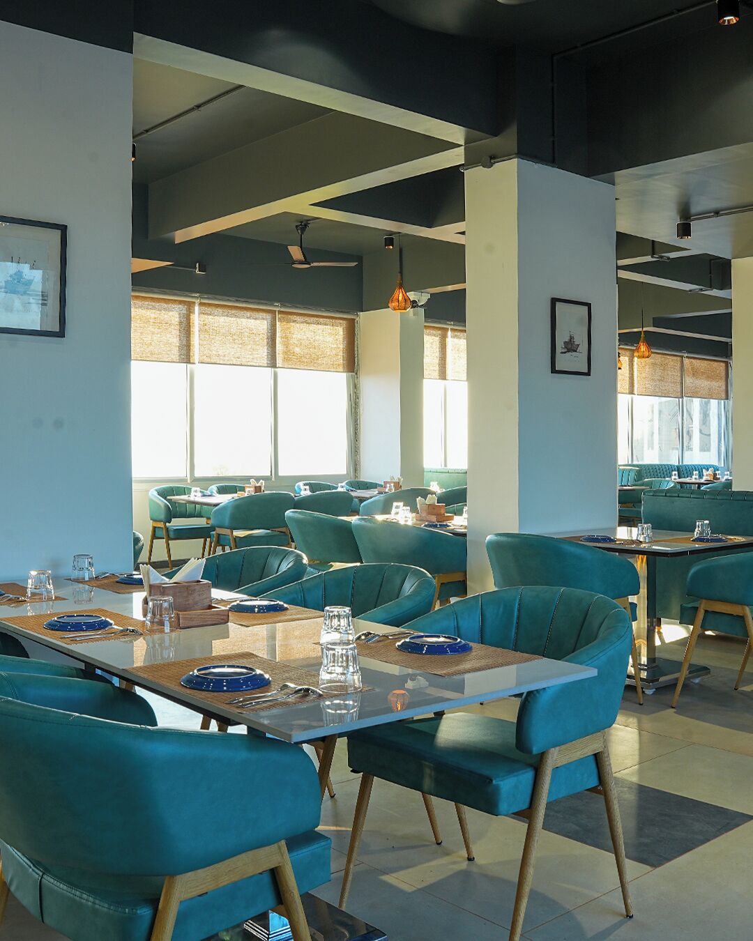 A restaurant with teal chairs and tables, large windows, and hanging pendant lights.