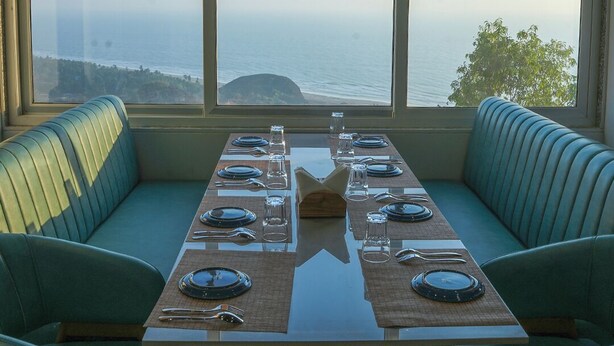 Comedor con una mesa preparada para cuatro, dos asientos tapizados en verde y vista al océano a través de amplias ventanas.