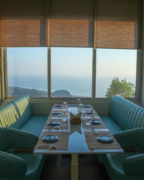 Uma área de refeições com mesa posta para quatro pessoas, dois assentos estofados verdes e vista para o oceano através de amplas janelas.