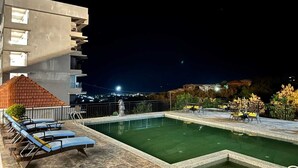 Una piscina en la azotea con sillas de descanso y una estatua, rodeada por una barandilla y vista nocturna de la ciudad.