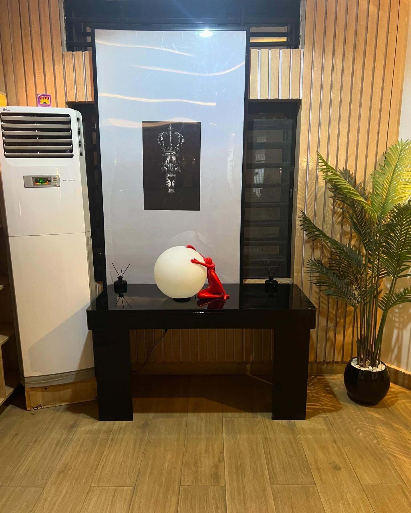A room with a black-framed mirror, a white air conditioner, a framed artwork, a red sculpture, and a potted plant.