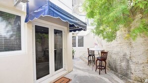 Um pequeno pátio com mesa e cadeiras, um toldo azul e um tapete de boas-vindas.