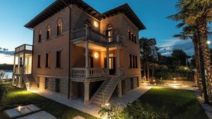 A two-story house with a balcony, a staircase, and a well-lit garden at dusk. - Ca' delle Contesse   Elegant 2BR apartment in Liberty villa   spectacular view! (Lido)