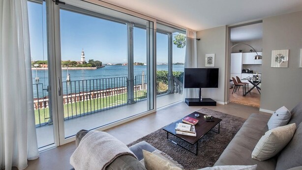 A modern living room with a large sliding glass door leading to a balcony overlooking a body of water and a lighthouse. - Ca' delle Contesse   Elegant 2BR apartment in Liberty villa   spectacular view! (Lido)