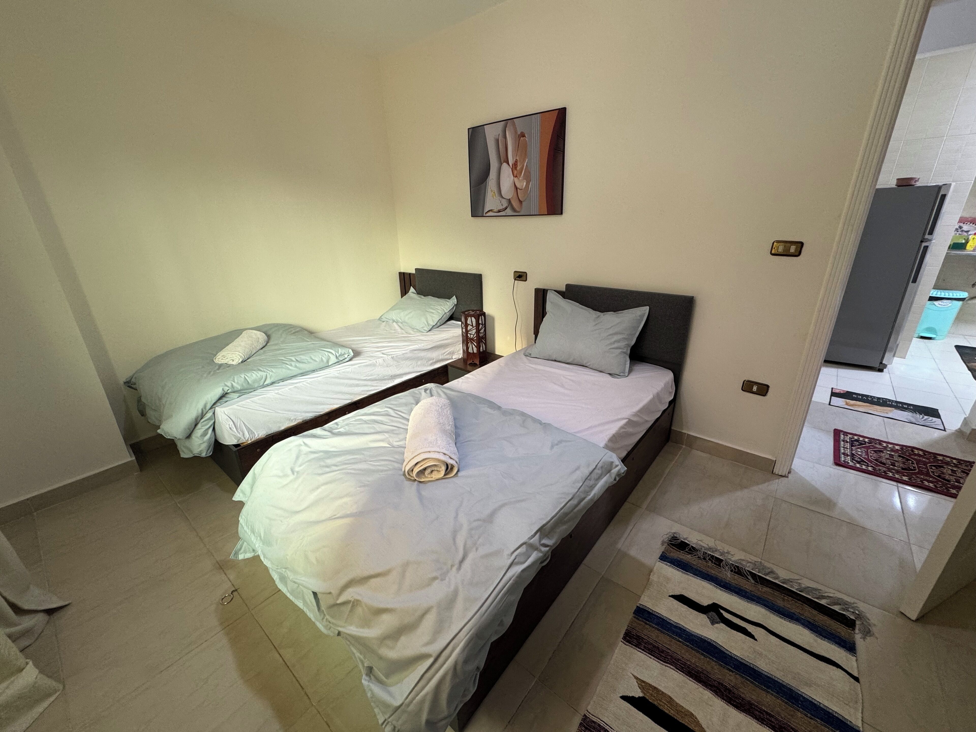 A hotel room with two beds, a painting on the wall, and a small table with a lamp.