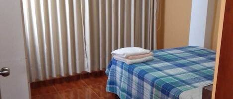 A small, tidy room with a bed, a wooden floor, and a window with curtains.