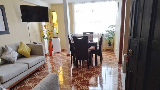A living room with a sofa, a dining table with chairs, a TV mounted on the wall, and a tiled floor.