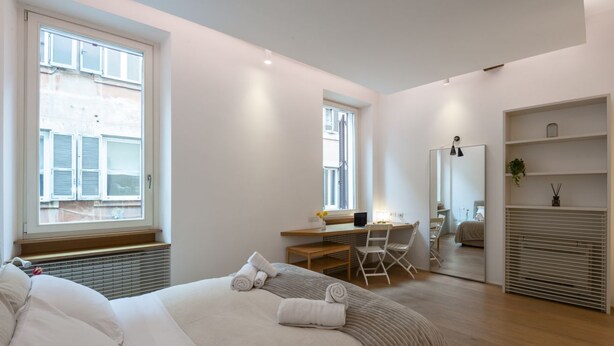 A modern bedroom with a large bed, a desk, and a chair. There is a window with blinds, a radiator, and a mirror on the wall.